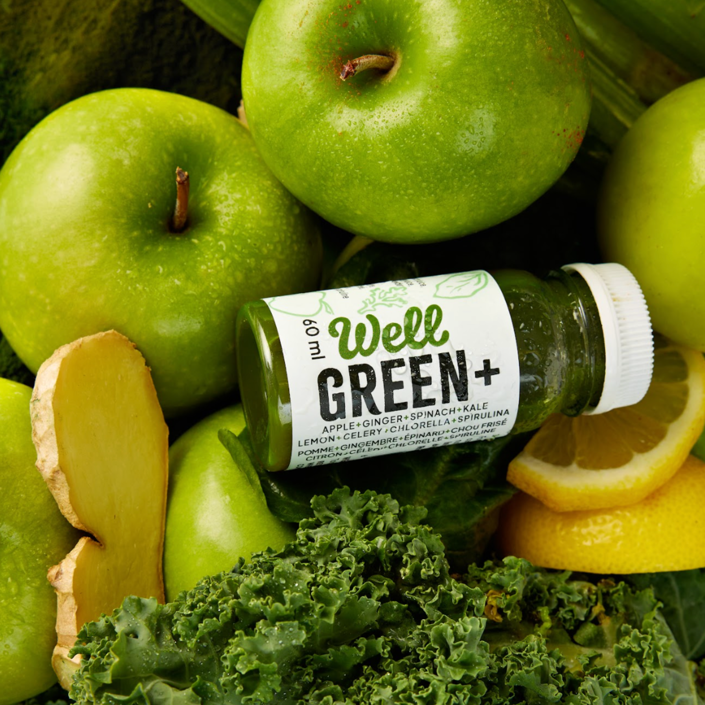 Bottle of Shot Well Green+ surrounded by ingredients: apple, ginger, spinach, kale, lemon, celery, chlorella, spirulina