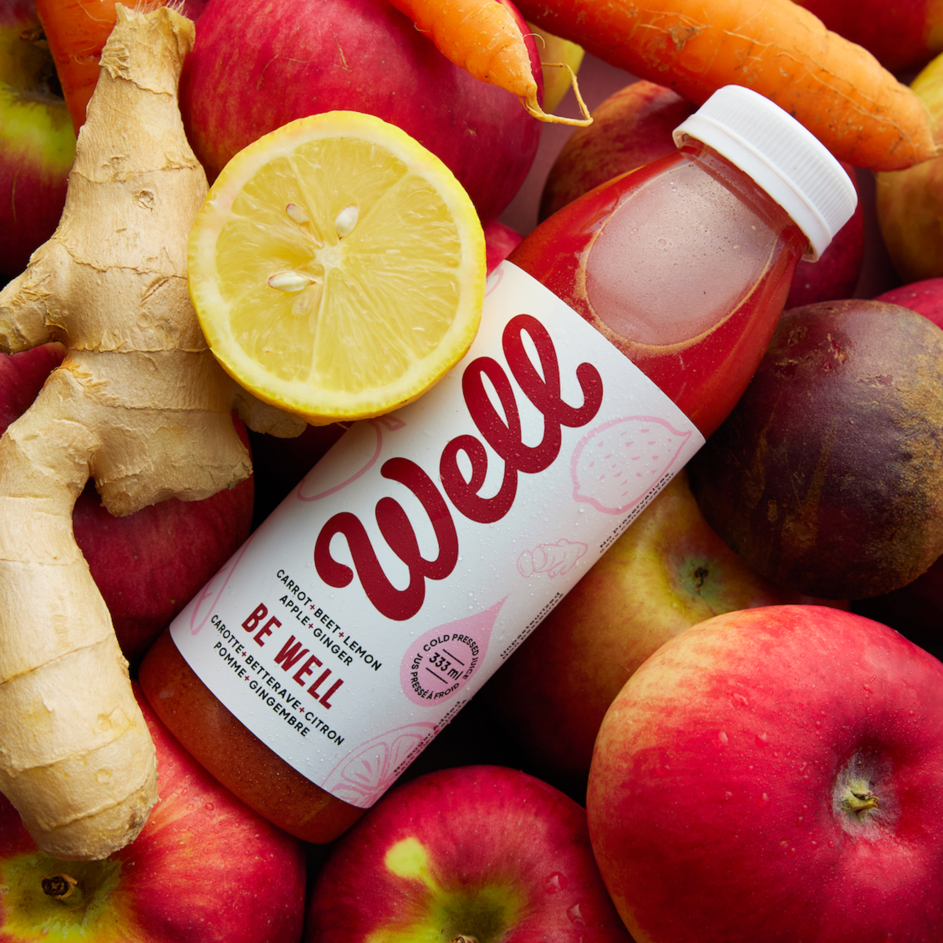 Bottle of 'Be Well' juice surrounded by fruits and vegetables on a colorful background: carrot, beet, lemon, apple, ginger